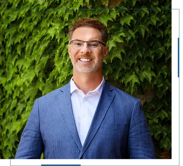 Man in blue suit smiling against leafy background.